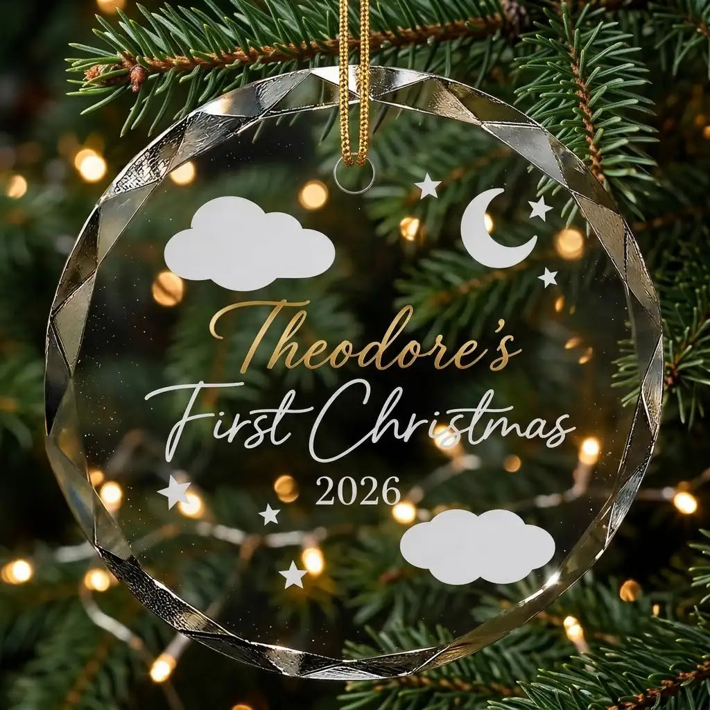 Personalized clear glass baby first christmas ornament displayed hanging on Christmas tree branch with warm sparkling holiday lights background