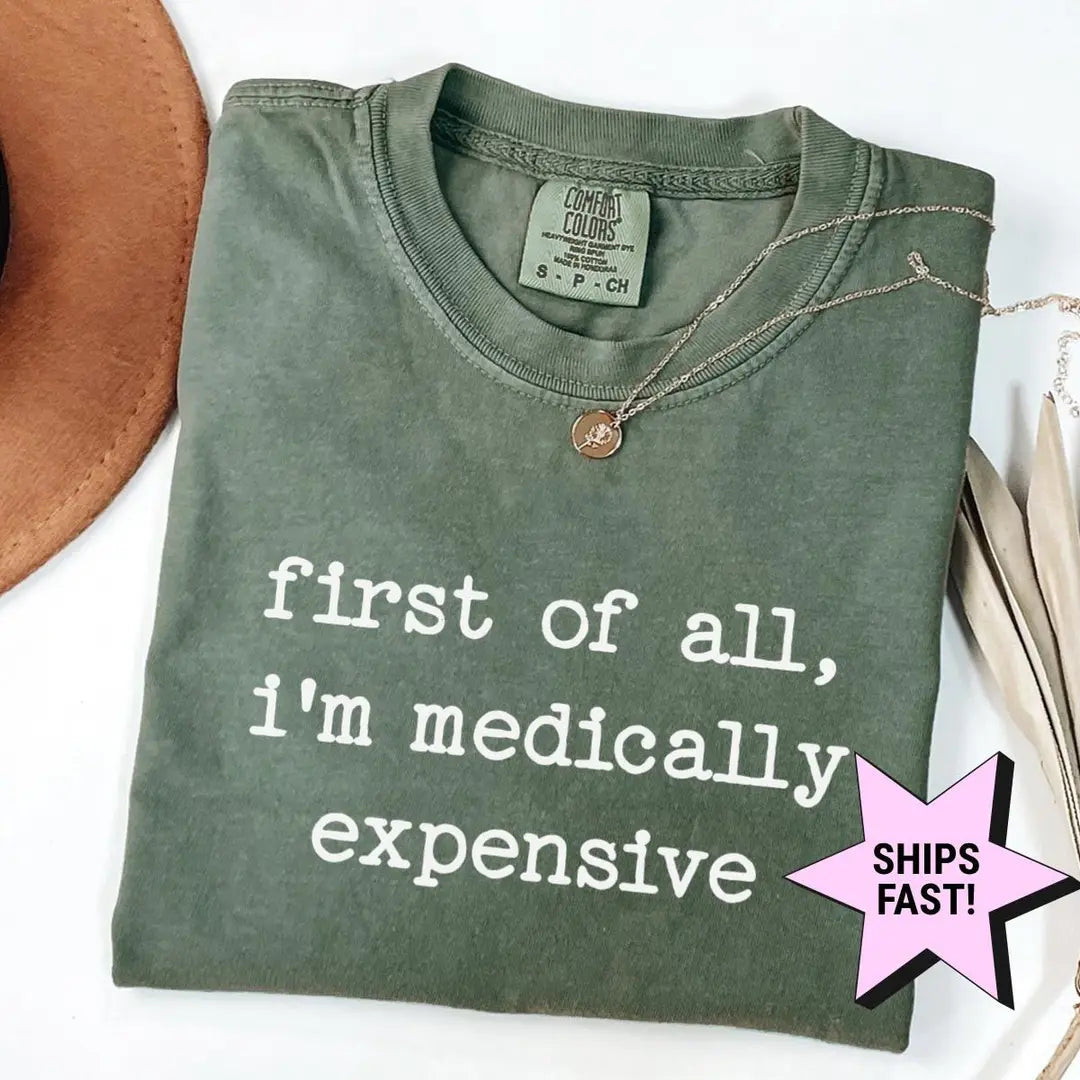 Folded Moss green Comfort Colors tee printed first of all, i'm medically expensive in white typewriter letters