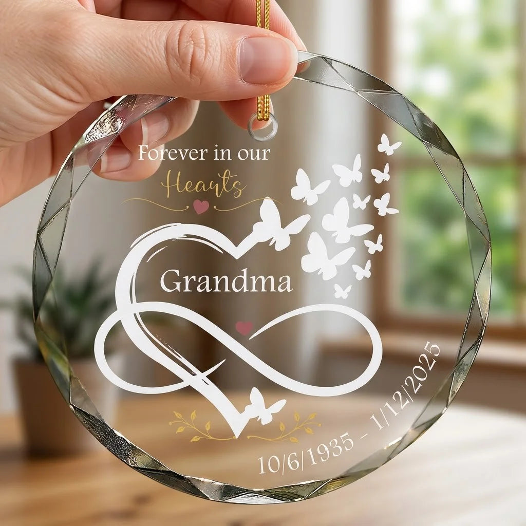 Hand holding a transparent faceted glass ornament with gold string loop showing printed infinity symbol merged heart with flying butterflies design and crisp white lettering in cozy indoor scene