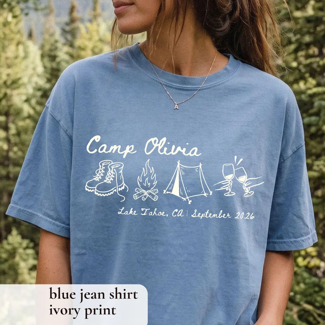 Woman in a Blue Jean Comfort Colors tee in a forest, ivory Camp Olivia script with hiking boots, campfire and tent