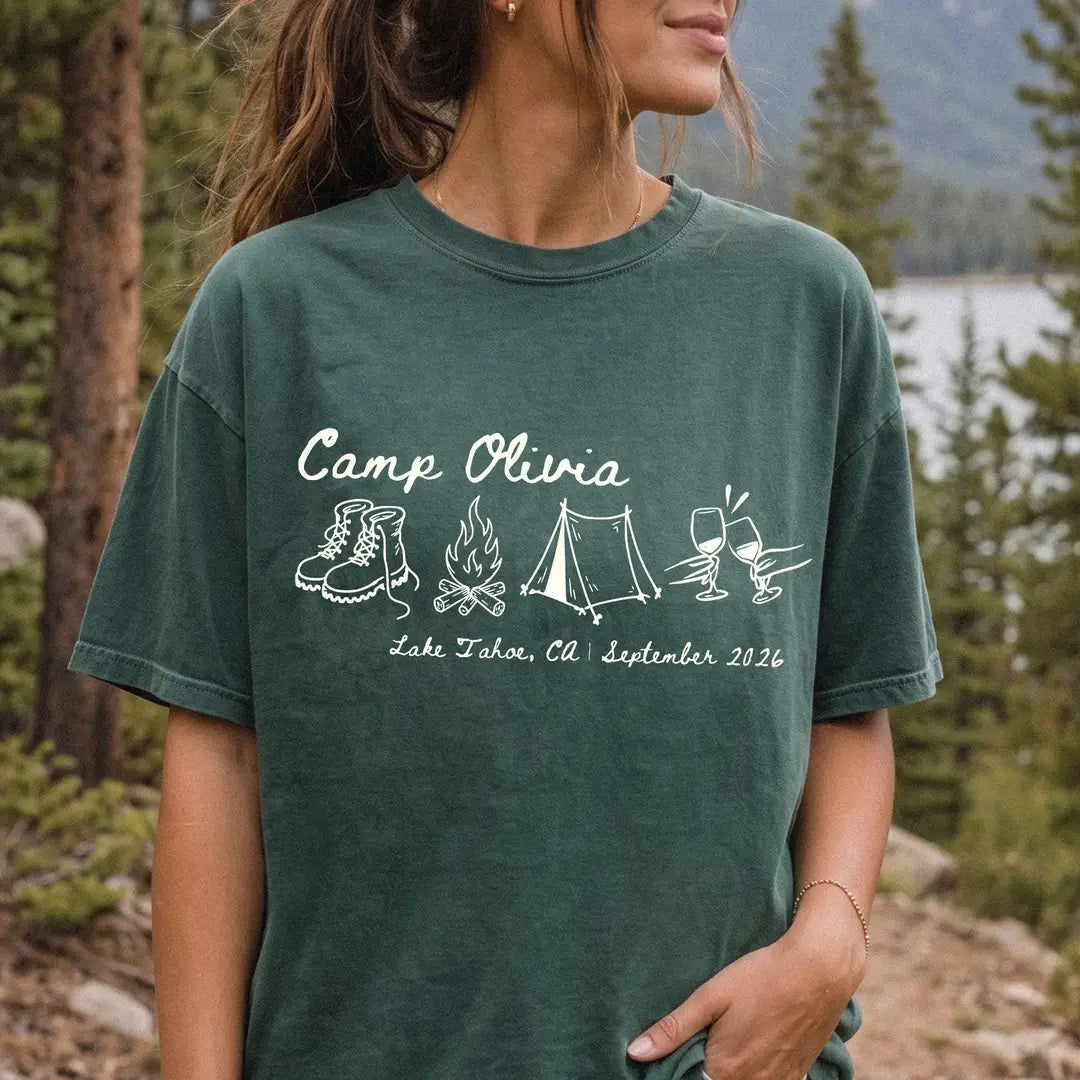 Woman in a Blue Spruce Comfort Colors tee in a pine forest, ivory Camp Olivia script with boots, campfire and tent