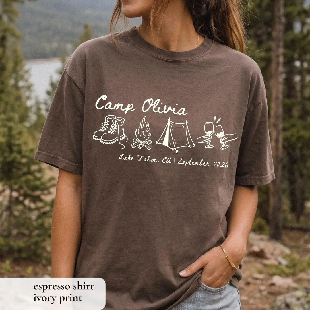 Woman in an Espresso brown Comfort Colors tee on a mountain trail, ivory Camp Olivia with boots, tent and wine toast