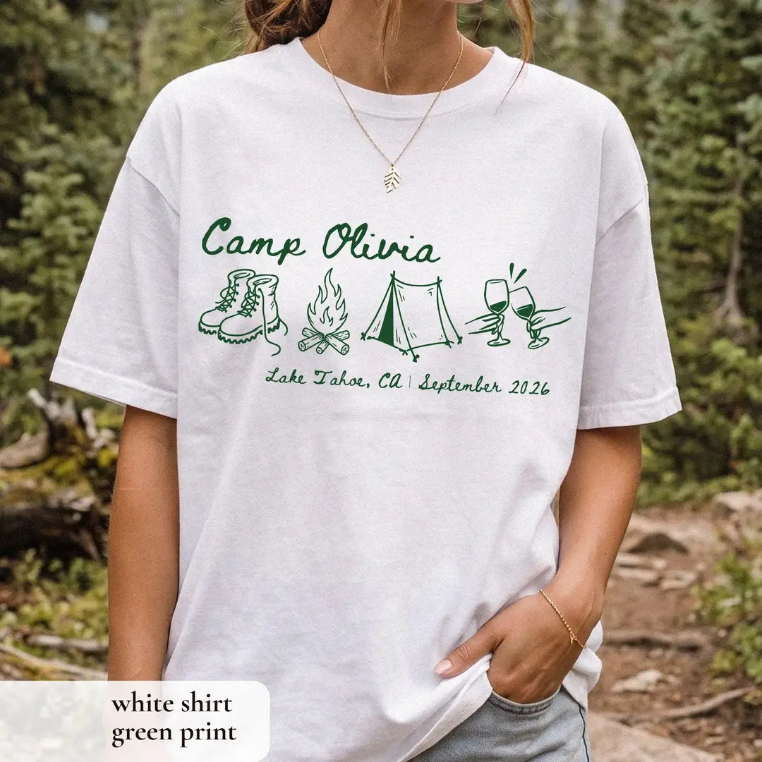 Woman in a White Comfort Colors tee among tall pines, green Camp Olivia script with boots, campfire, tent and wine