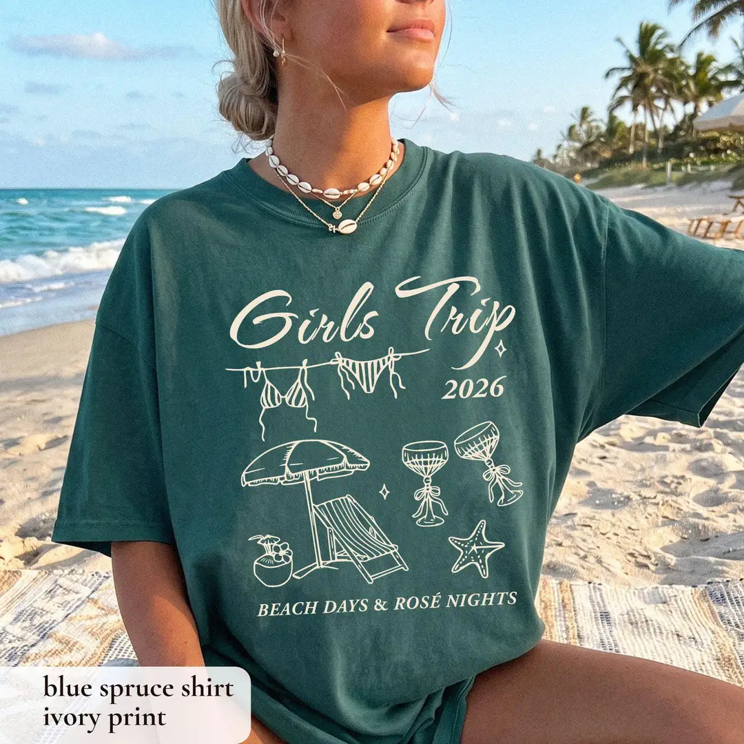 Woman in a Blue Spruce Comfort Colors tee on a beach, ivory girls trip 2026 with bikinis, umbrella and wine glasses