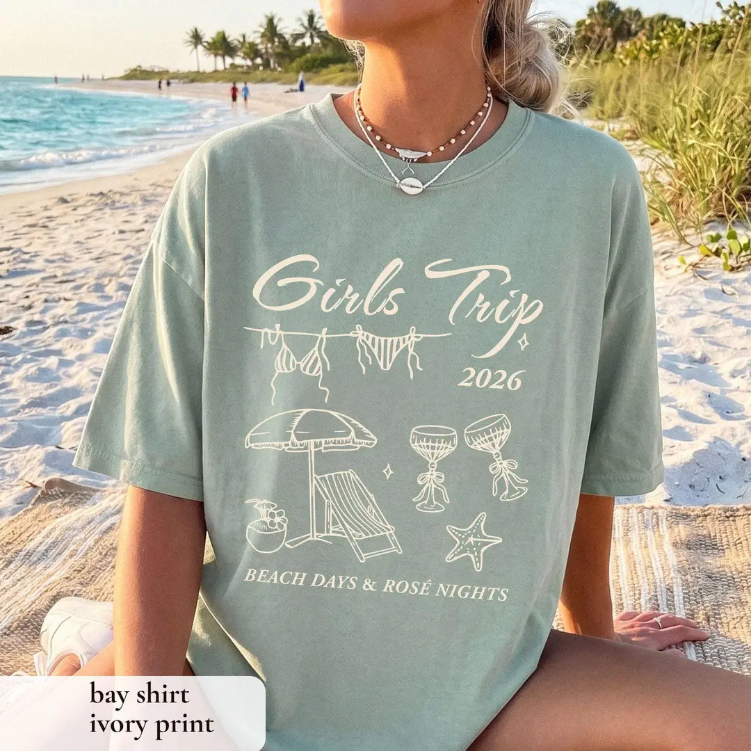 Woman in a Bay sage Comfort Colors tee on a sandy beach, ivory girls trip 2026 with umbrella, lounge chair and starfish