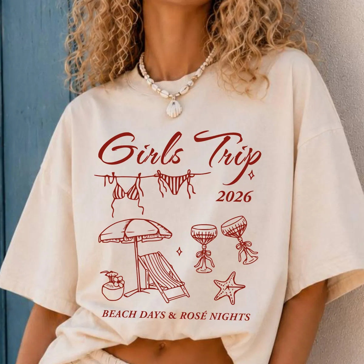 Woman in an Ivory Comfort Colors tee leaning on a blue wall, crimson girls trip 2026 script, bikinis and beach icons
