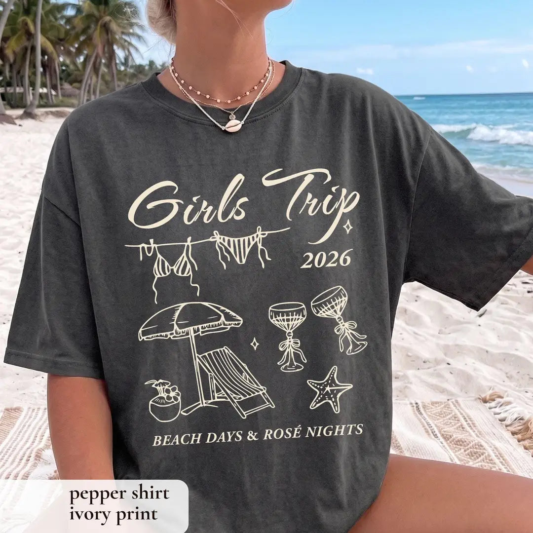 Woman in a Pepper Comfort Colors tee on a palm beach with ivory girls trip 2026 script and a bikini clothesline print