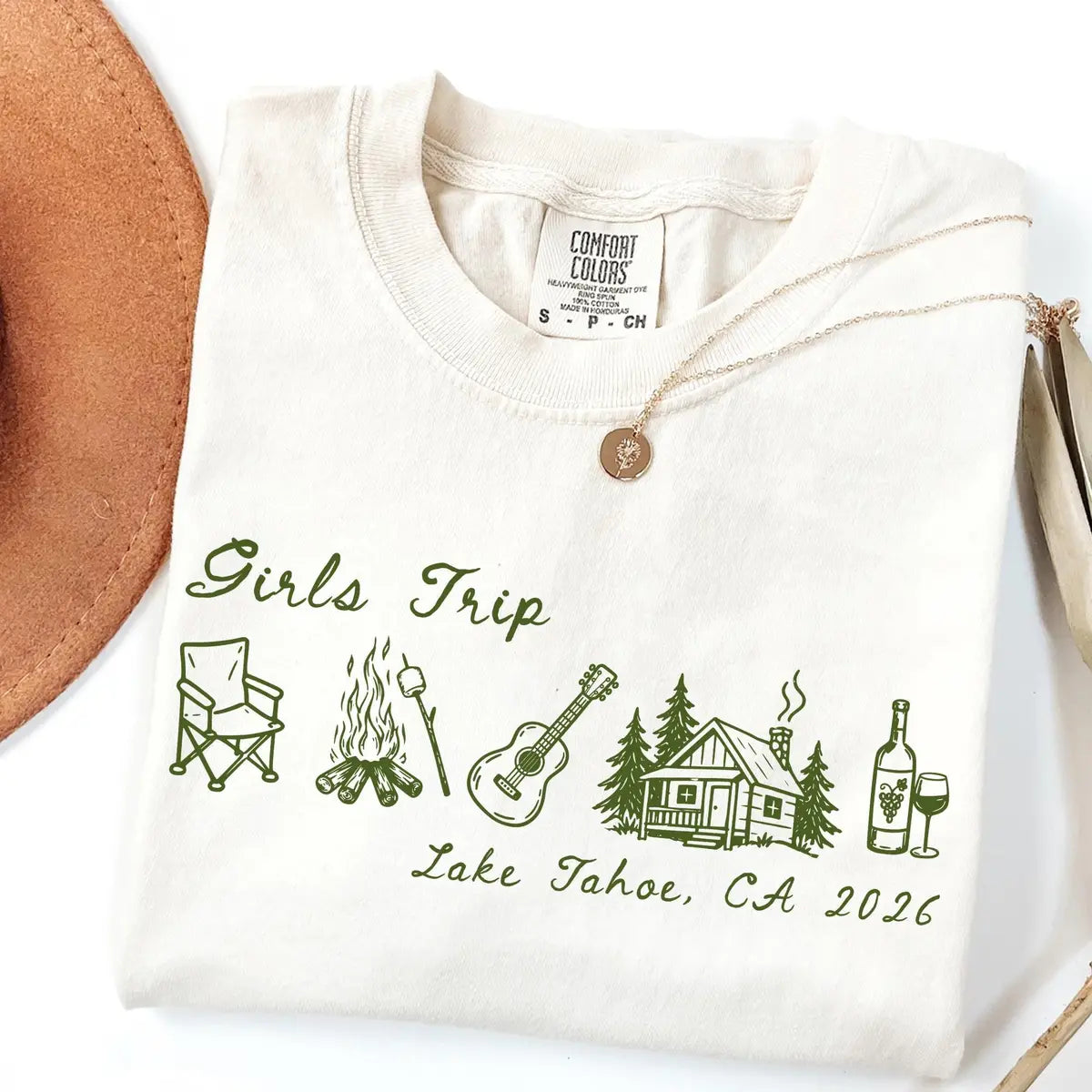 Folded Ivory Comfort Colors tee with green girls trip script, camp chair, campfire, guitar and cabin, tan hat beside