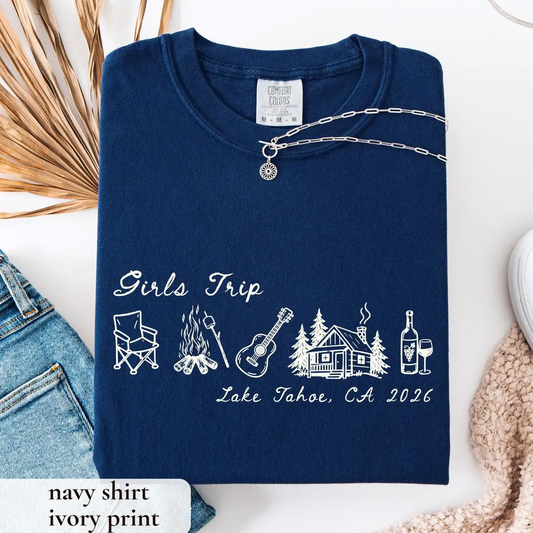 Folded True Navy Comfort Colors tee with ivory girls trip script, camp chair, campfire, guitar and cabin, jeans beside