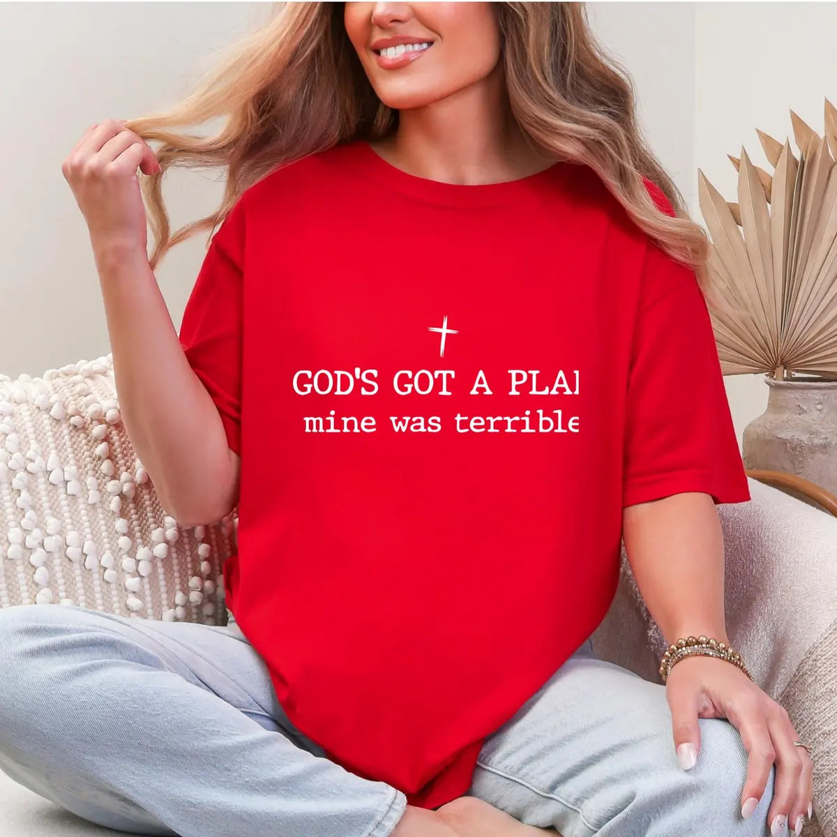Woman in a Red Comfort Colors tee sitting cross-legged, a small white cross above god's got a plan, mine was terrible