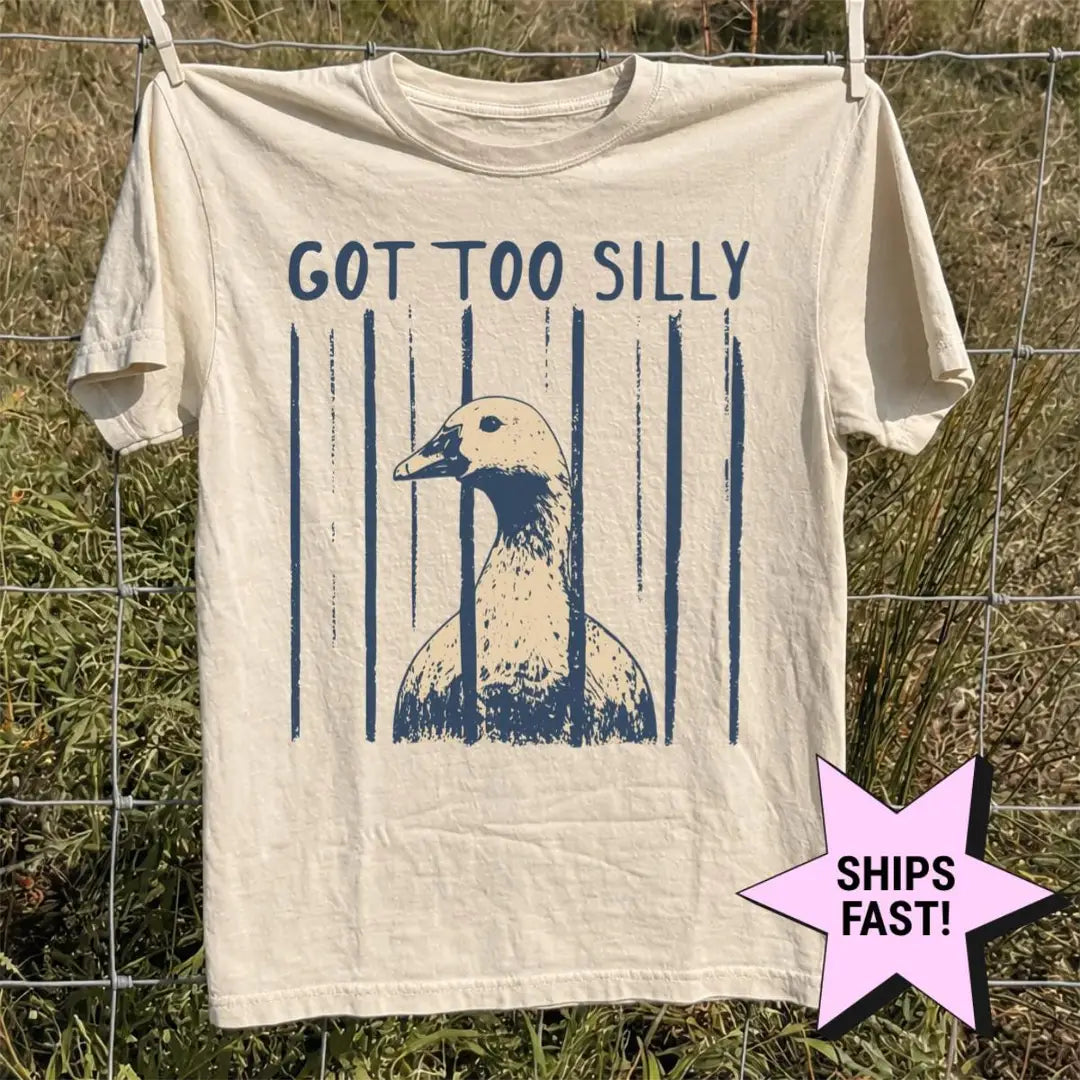 Ivory Comfort Colors tee hanging on a wire fence in tall grass, got too silly above a navy and tan goose head print