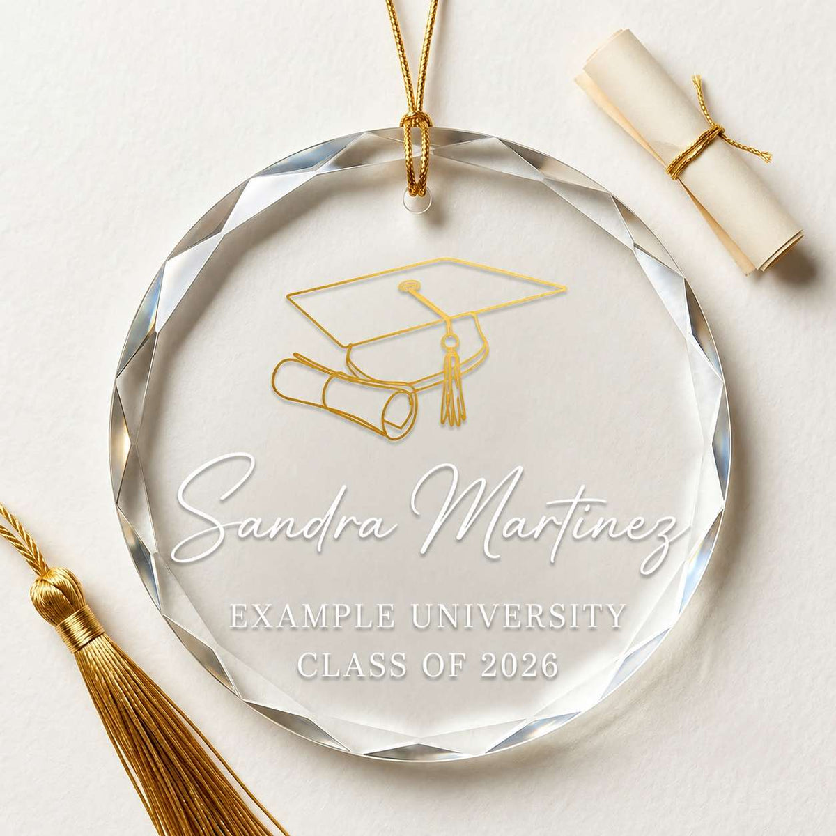 A round clear acrylic graduation ornament with a faceted edge, hanging from a gold string. The ornament is engraved with a gold graduation cap and scroll design, the text in white block text.
