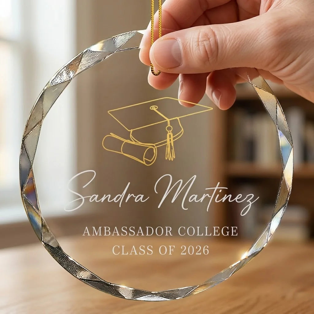 Hand holding a transparent faceted glass ornament with gold string loop showing printed graduation cap and diploma scroll design and crisp white lettering in cozy indoor scene
