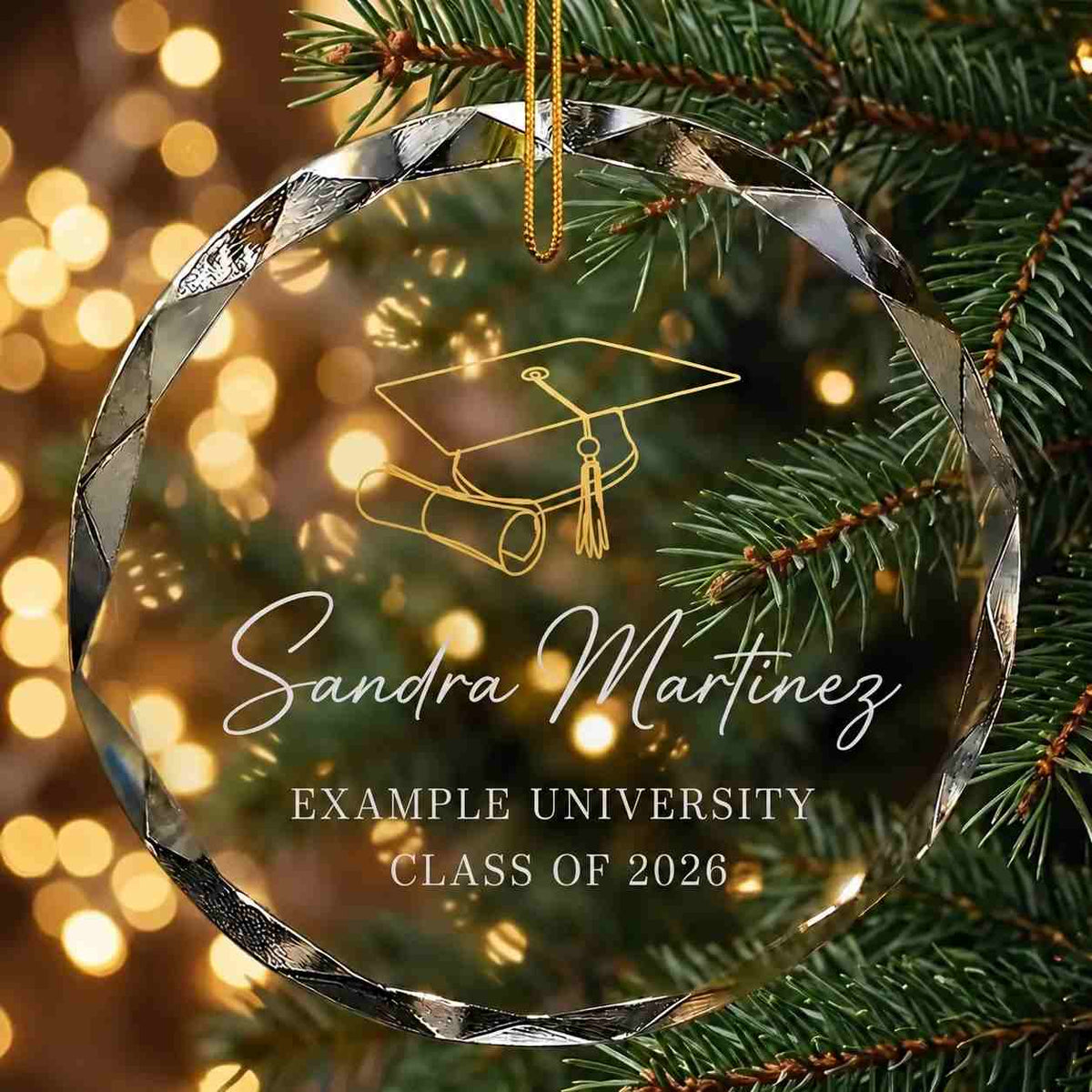 A personalized graduation ornament hanging on a Christmas tree branch. The faceted clear ornament features a gold laser-engraved graduation cap and diploma design, with the name printed in white.