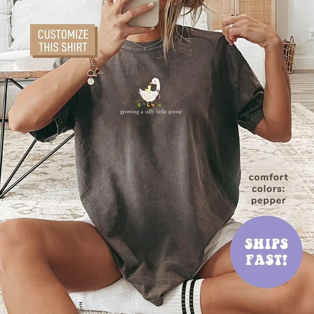 Woman in a Pepper Comfort Colors tee taking a mirror selfie, a small goose with ducklings above growing a silly little goose