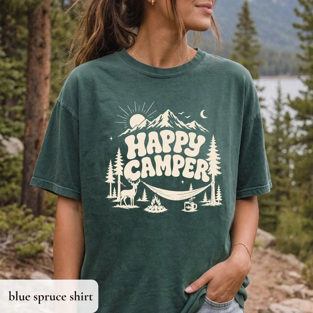 Woman in a Blue Spruce Comfort Colors tee on a forest trail, ivory happy camper lettering, hammock and campfire
