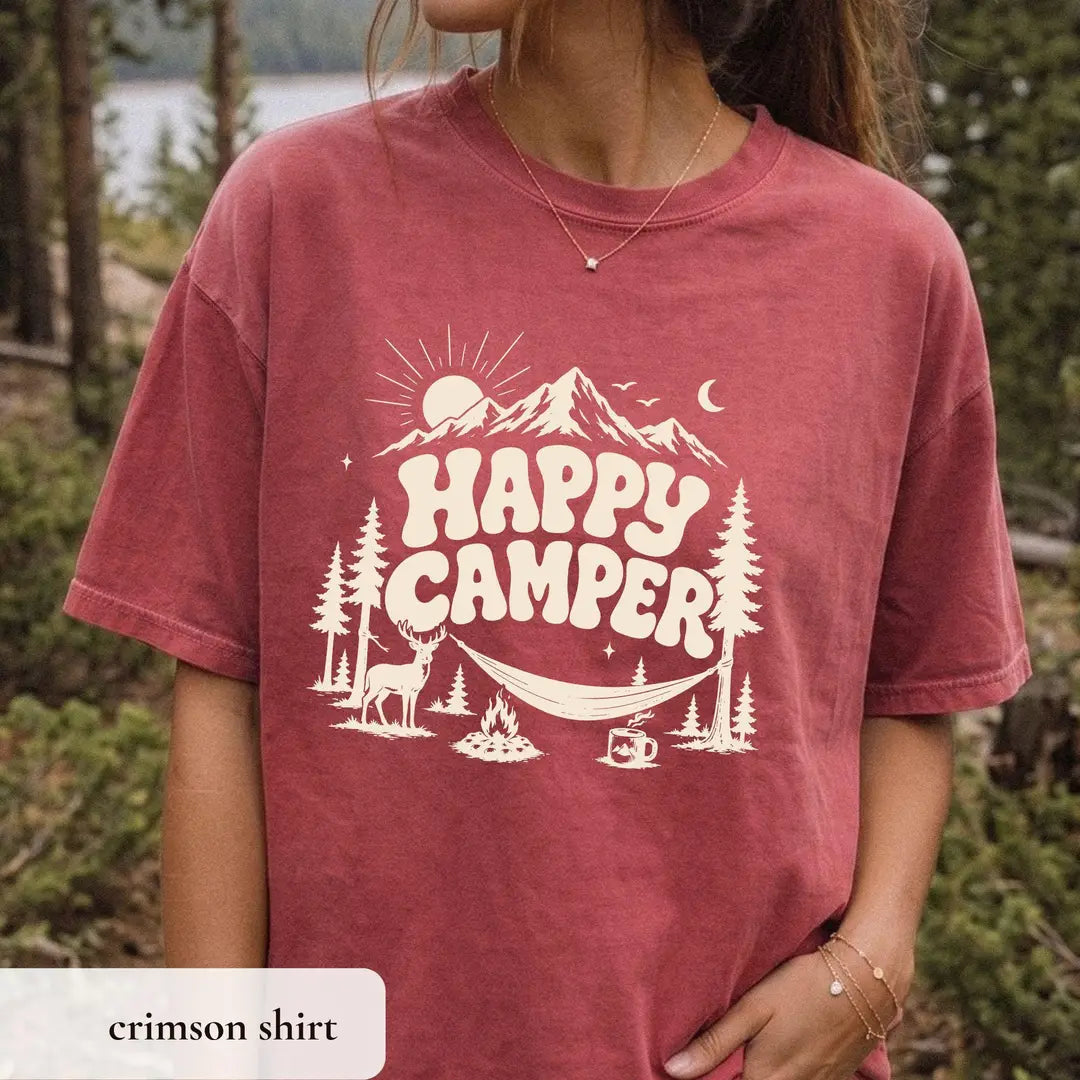 Woman in a Crimson Comfort Colors tee among pines, ivory happy camper lettering with a mountain, sun and hammock
