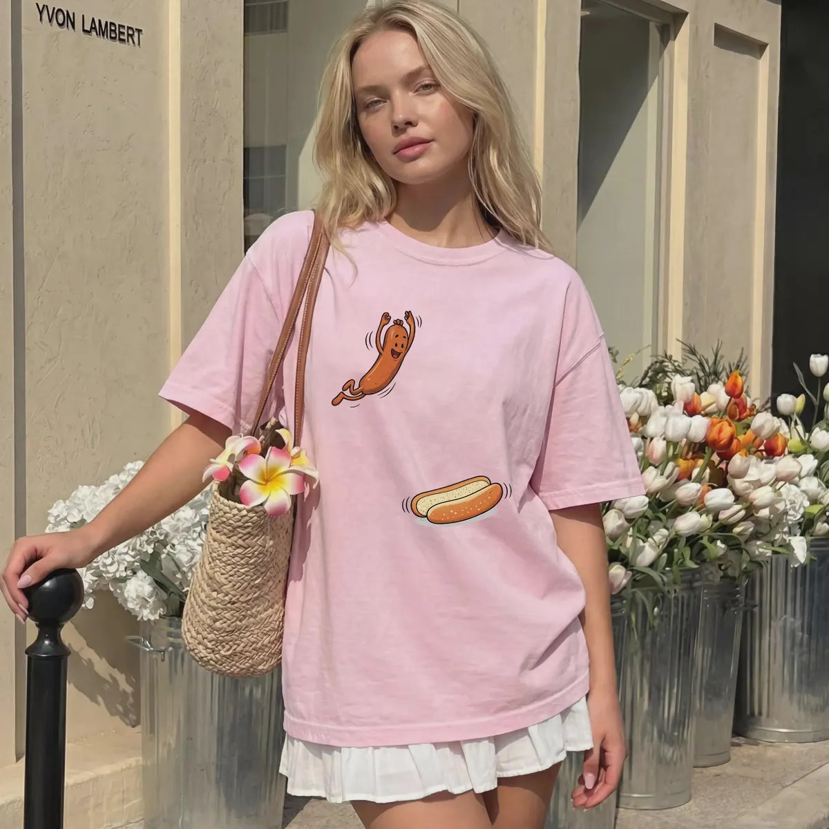 Blossom pink Comfort Colors tee with a cartoon hot dog jumping toward a bun, worn outside a shop with a woven bag