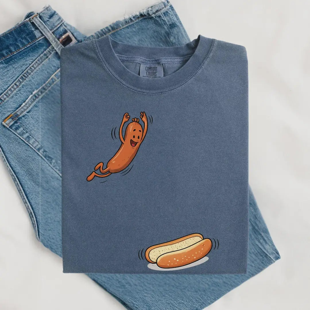 Folded Blue Jean Comfort Colors tee on denim with a cartoon hot dog leaping toward a bun near the hem