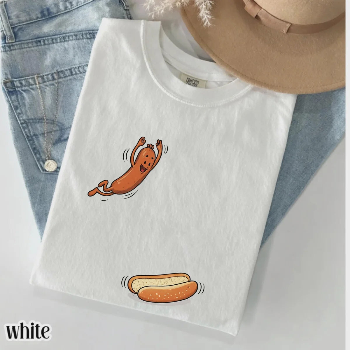 White Comfort Colors tee folded on jeans with a cartoon hot dog jumping toward a bun, styled with a straw hat