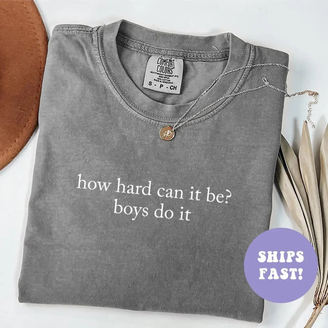 Folded Pepper Comfort Colors tee with how hard can it be? boys do it in white serif, beside a brown hat