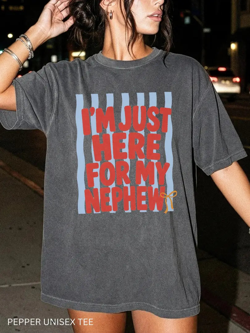 Woman in a Pepper Comfort Colors tee on a night street, red I'm just here for my nephew text on a blue striped panel