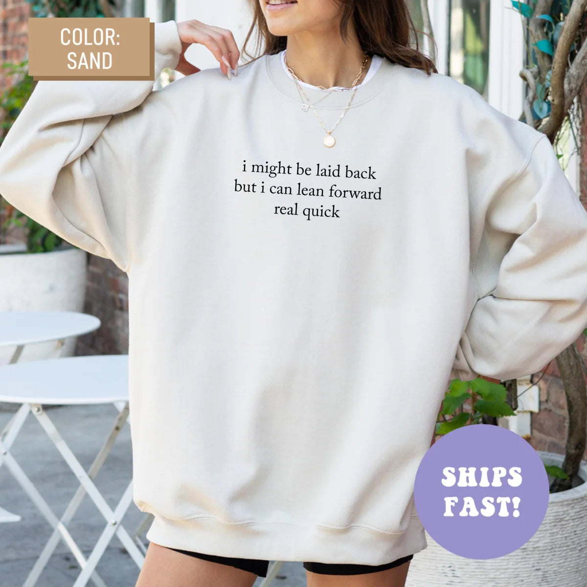 Woman in an oversized Sand sweatshirt, hands at her shoulders, dark serif quote, I might be laid back but I can lean forward