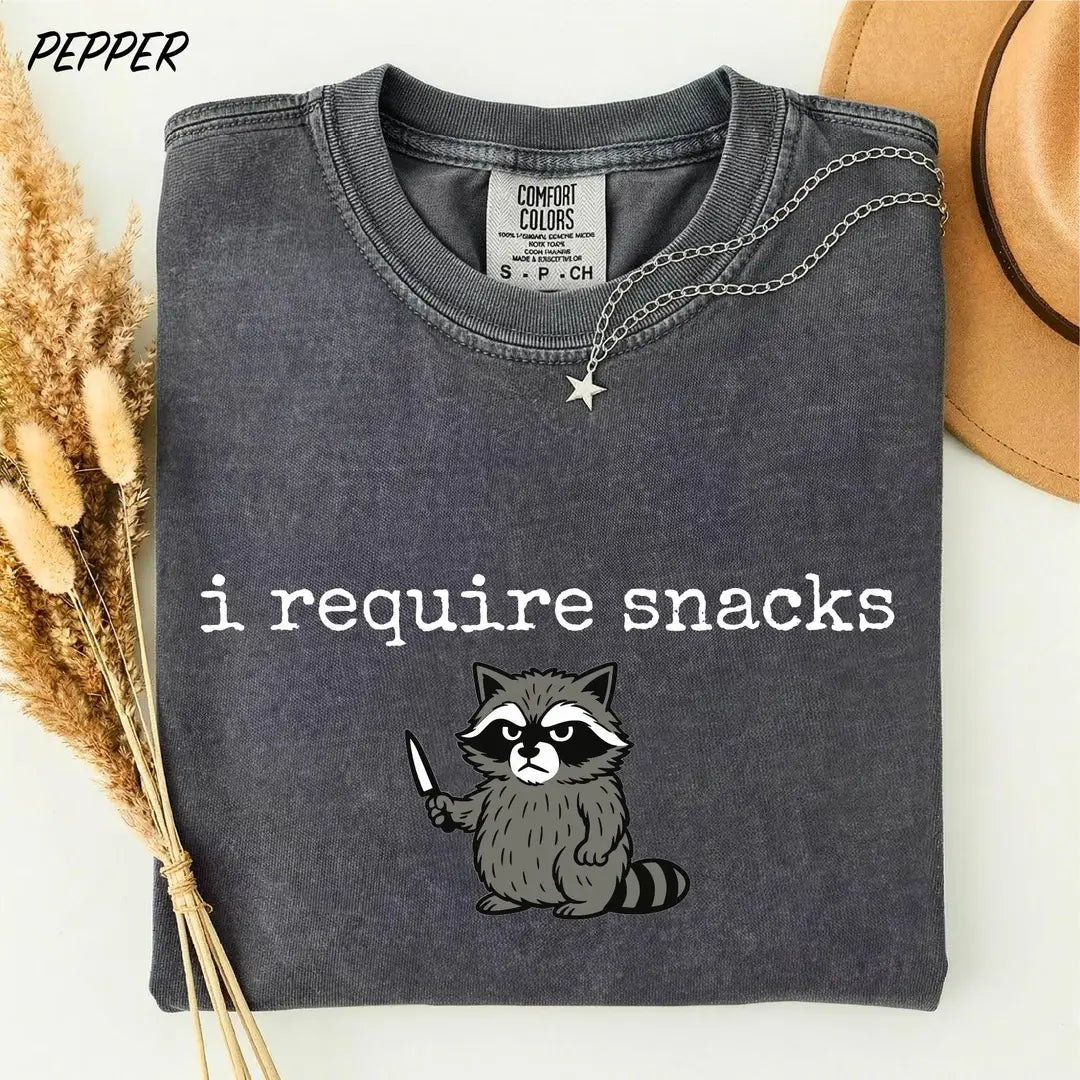 Pepper Comfort Colors 1717 tee with white 'I require snacks' text and a gray raccoon holding a small knife.