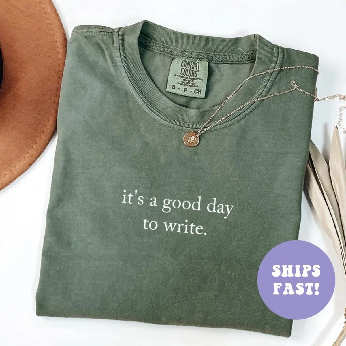 Folded Moss green Comfort Colors tee with it's a good day to write in white serif, dried palm leaves nearby
