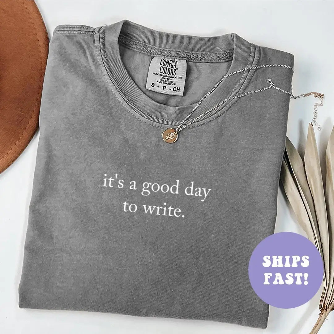 Folded Pepper Comfort Colors tee with it's a good day to write in white serif, necklace and brown hat beside it