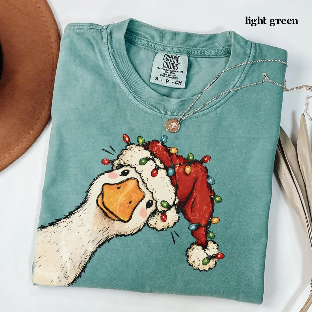 Folded Light Green Comfort Colors tee with a Santa hat goose and colorful bulbs, laid on a white surface