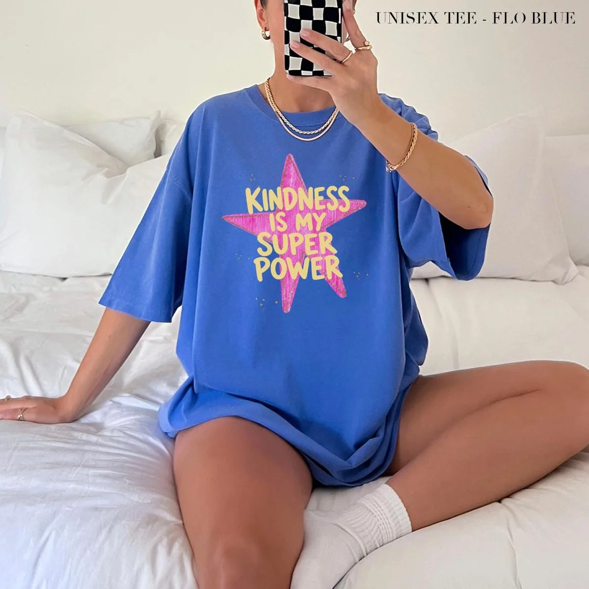 Woman in a Flo Blue Comfort Colors tee taking a mirror selfie on a bed, yellow kindness lettering on a pink star