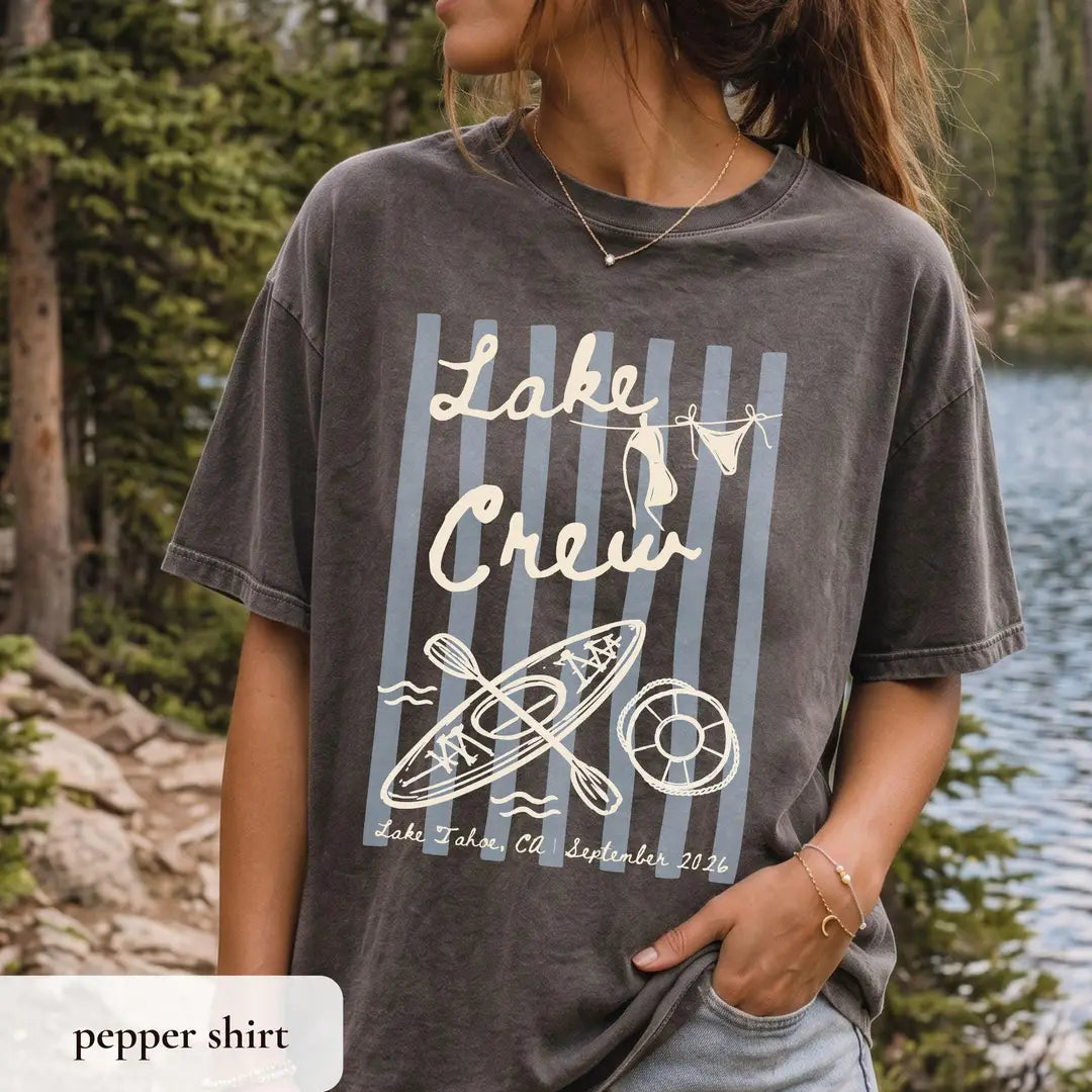 Woman in a Pepper Comfort Colors tee by a mountain lake, ivory lake crew script over stripes with a kayak and life ring