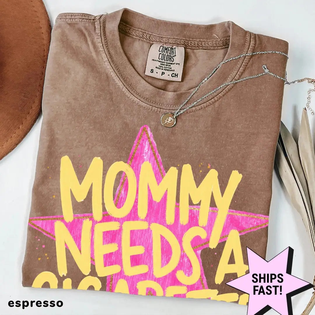 Folded Espresso brown Comfort Colors tee with big yellow MOMMY NEEDS A lettering over a pink star, necklace and hat nearby