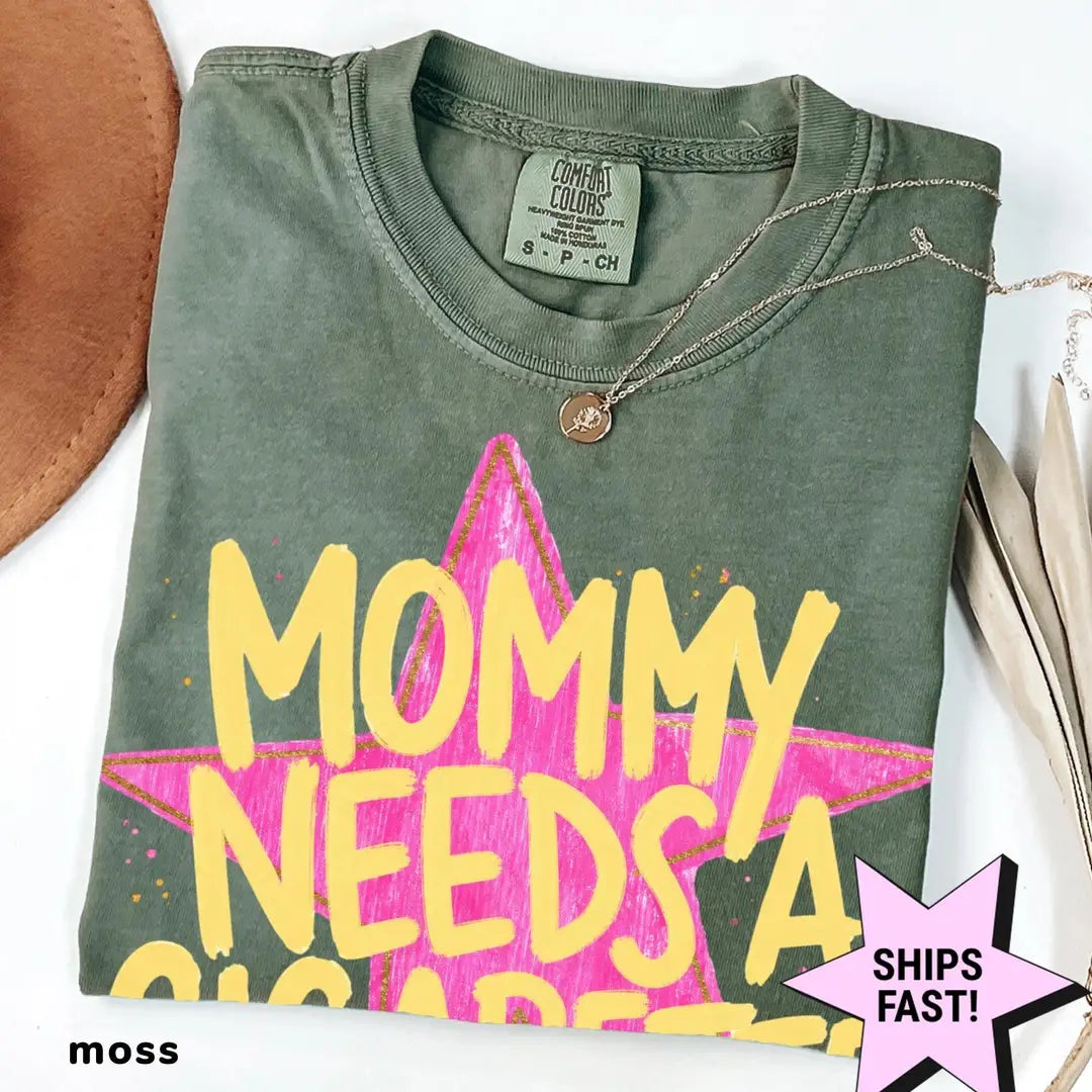 Folded Moss green Comfort Colors tee with large yellow MOMMY NEEDS A lettering on a pink brush star, necklace on the collar
