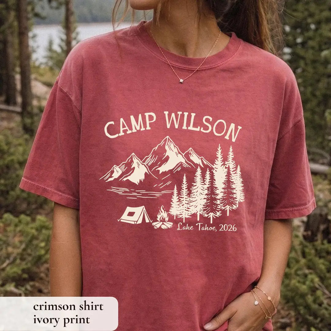 Woman in a Crimson Comfort Colors tee among pines, ivory Camp Wilson over a mountain range, lake, tent and campfire