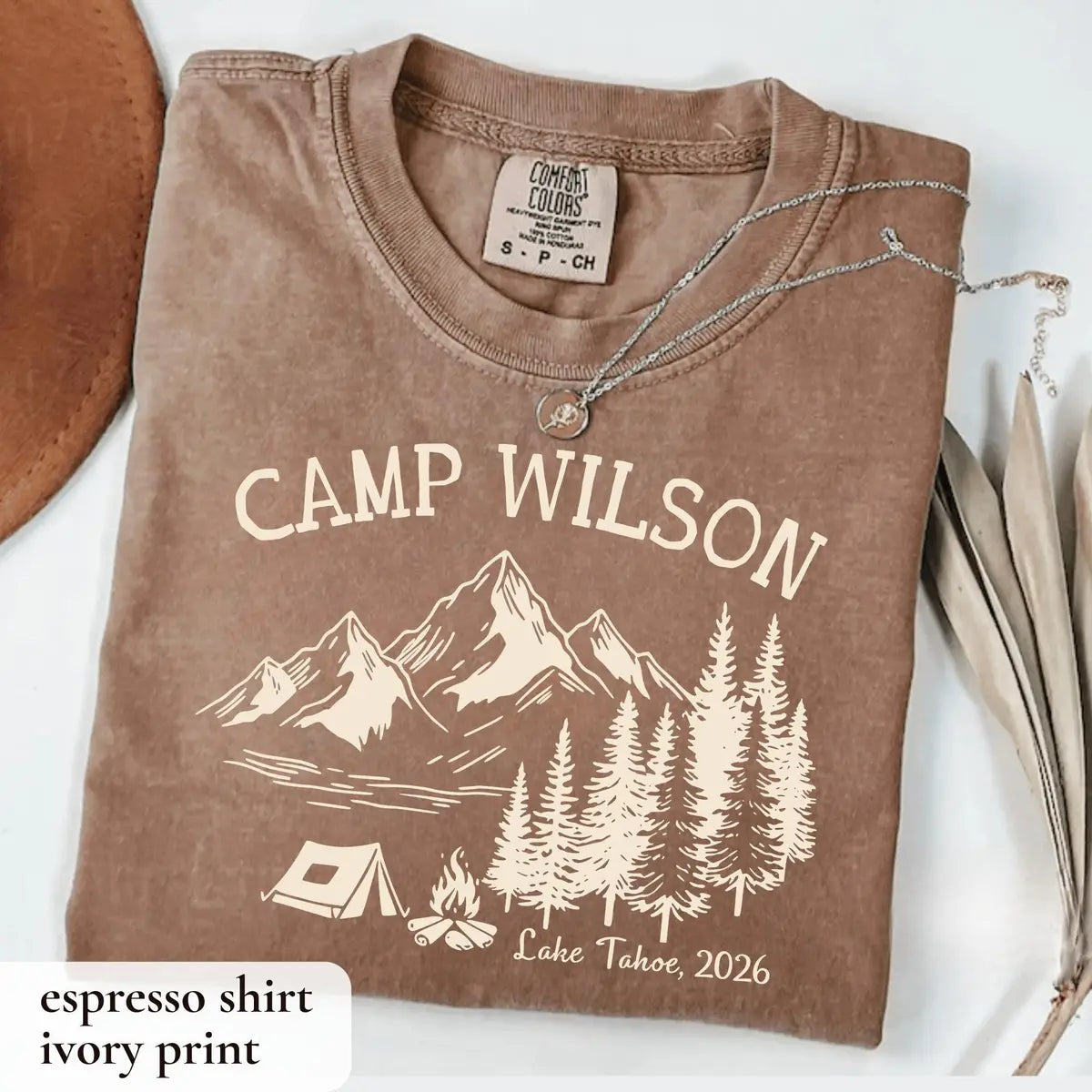 Folded Espresso brown Comfort Colors tee beside a tan hat, ivory Camp Wilson with mountains, pines, tent and Lake Tahoe, 2026