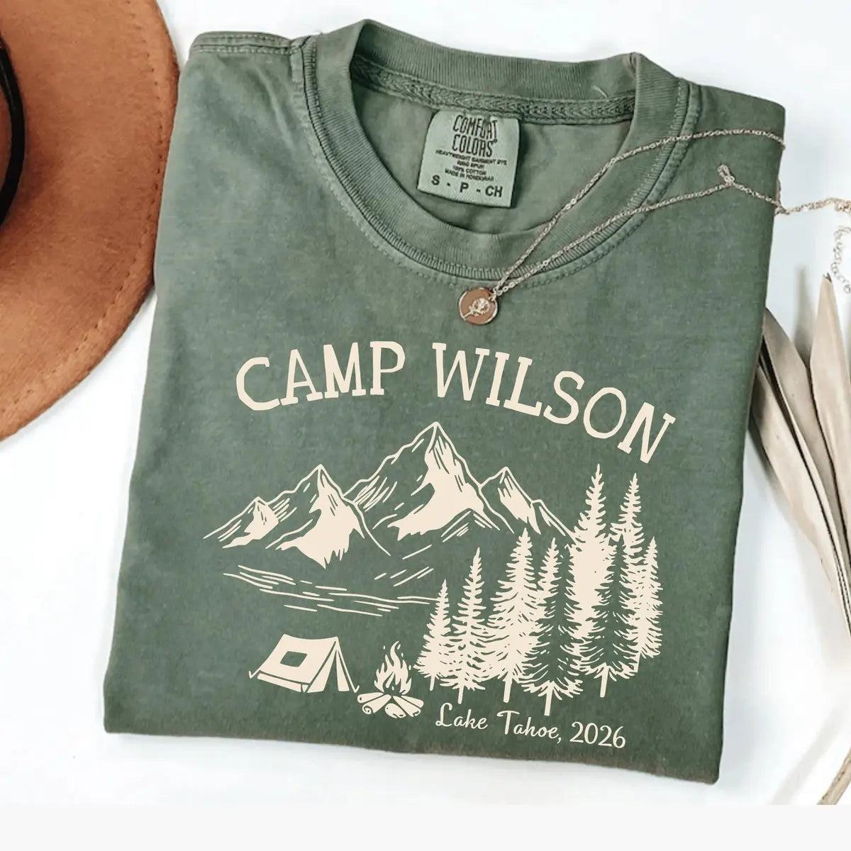 Folded Moss green Comfort Colors tee with ivory Camp Wilson above mountains, pines, a tent and campfire, Lake Tahoe, 2026