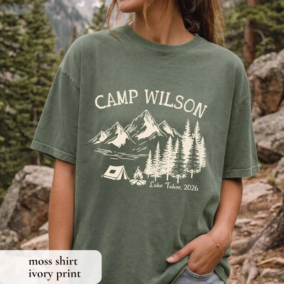 Woman in a Moss green Comfort Colors tee among rocks, hand in her pocket, ivory Camp Wilson over mountains, pines and tent