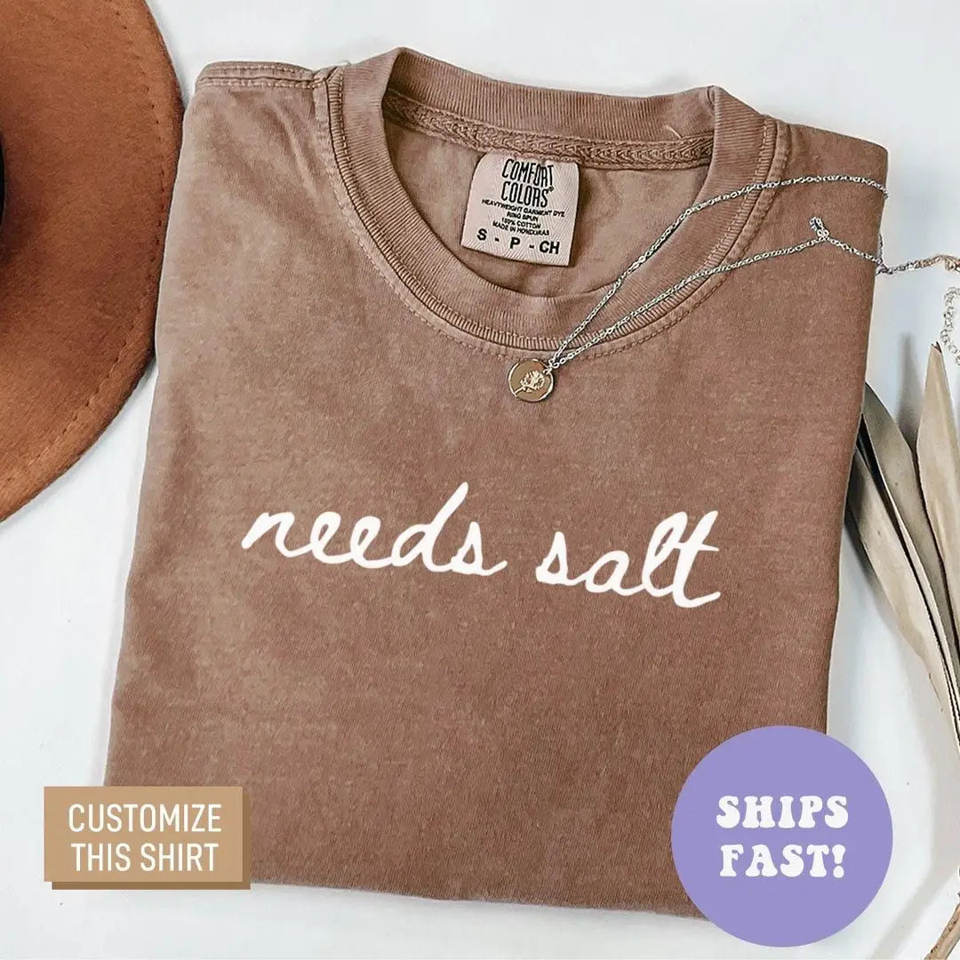 Folded Espresso brown Comfort Colors tee with needs salt in light cursive lettering, necklace at the neckline