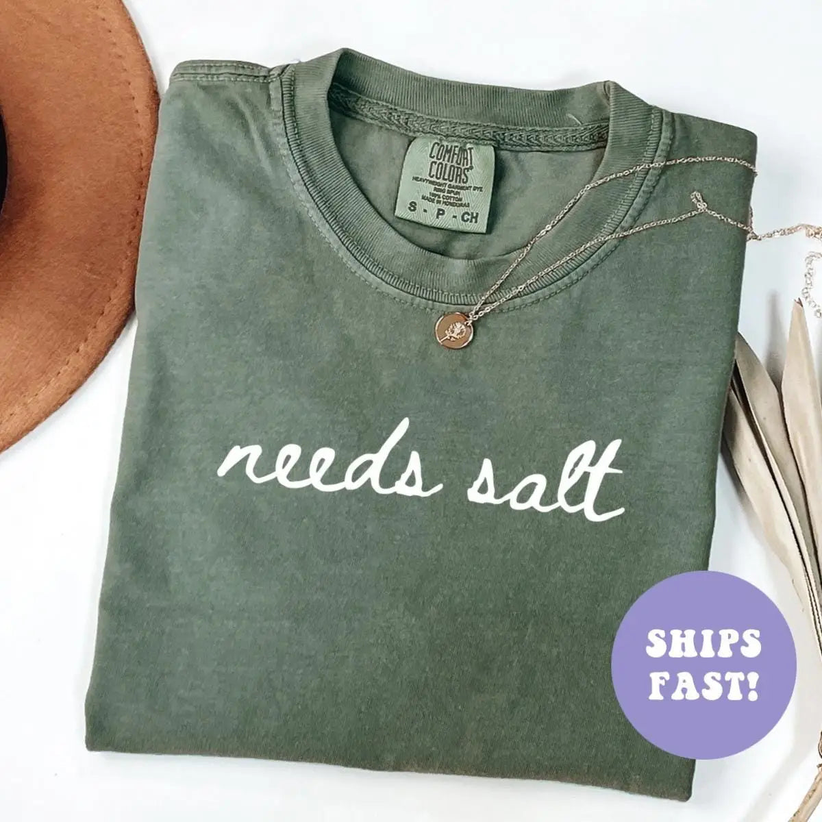 Folded Moss green Comfort Colors tee printed needs salt in lowercase white script, with a necklace on the collar