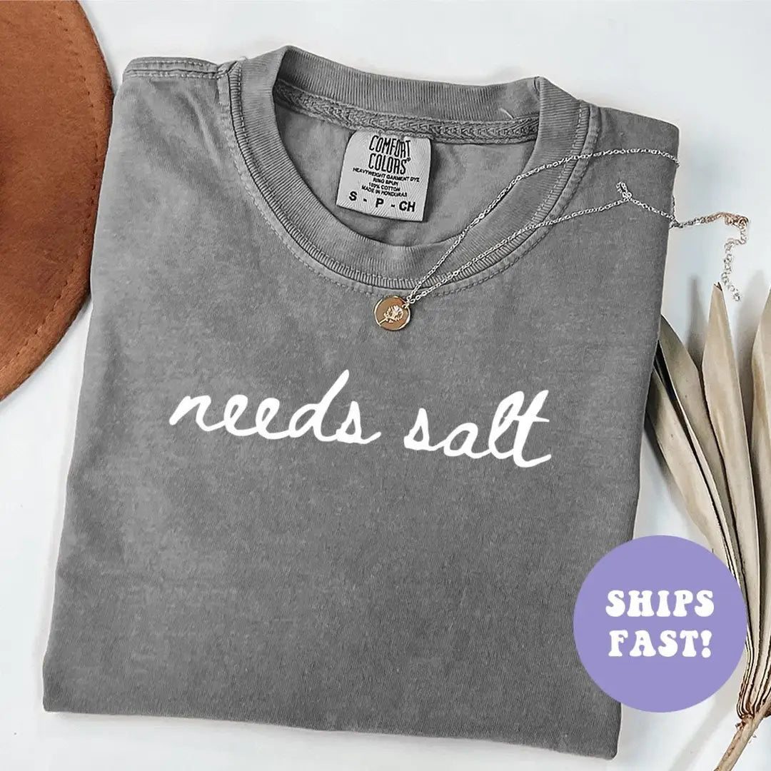 Folded Pepper Comfort Colors tee with needs salt in white cursive script, a necklace at the collar