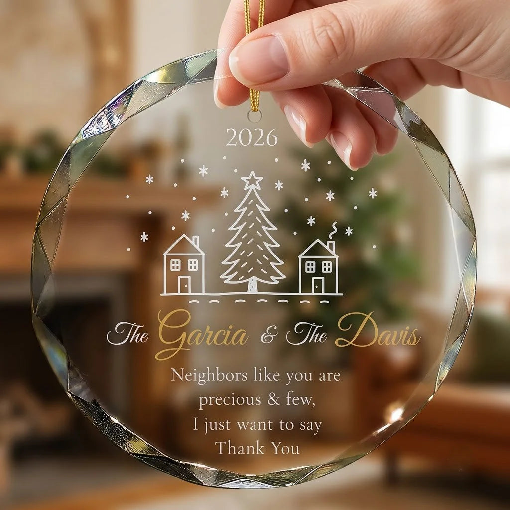 Hand holding a transparent faceted glass ornament with gold string loop showing printed two houses with pine tree in middle design and crisp white lettering in cozy indoor scene