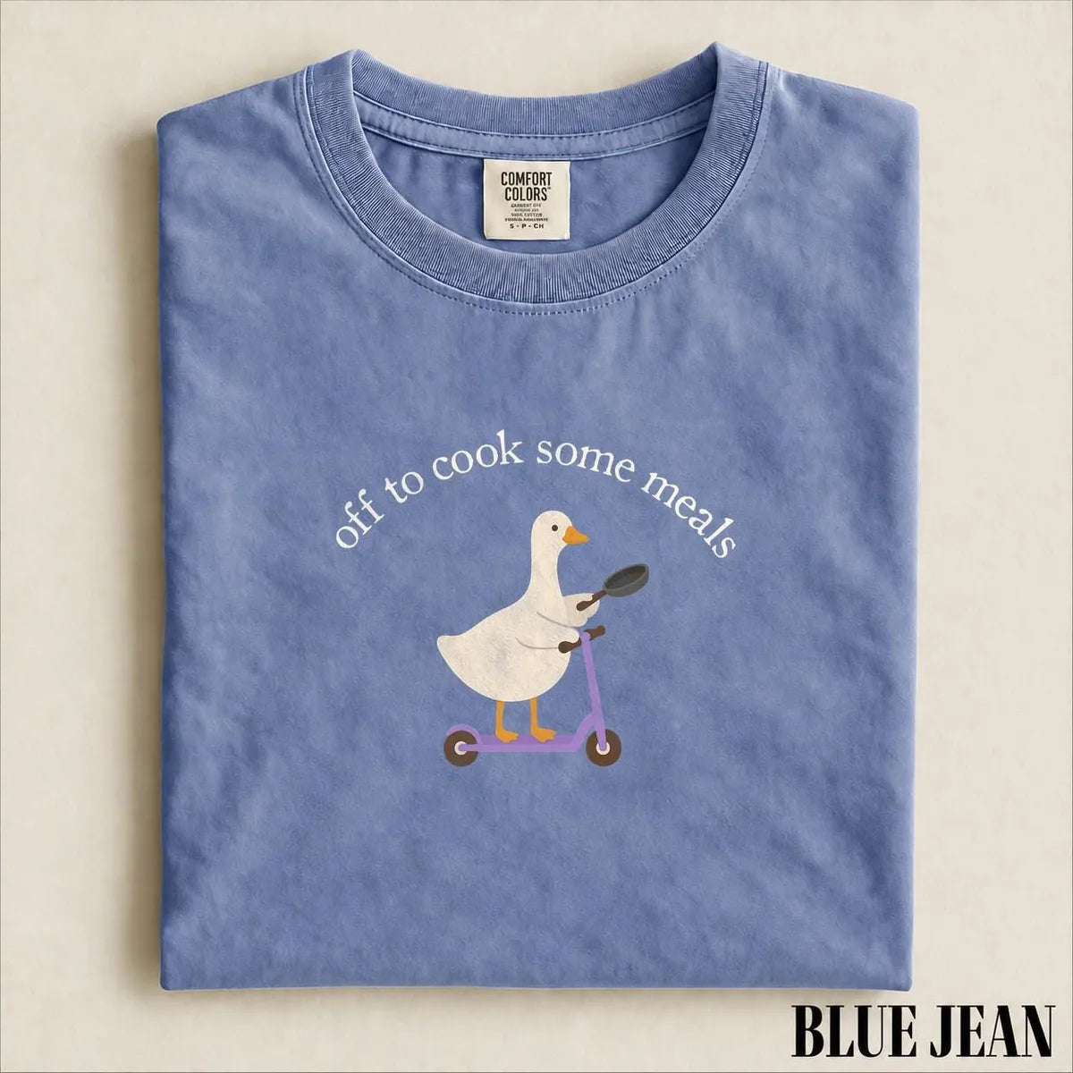 Folded Blue Jean Comfort Colors tee with a white goose, frying pan and purple scooter under the arched cook some meals text