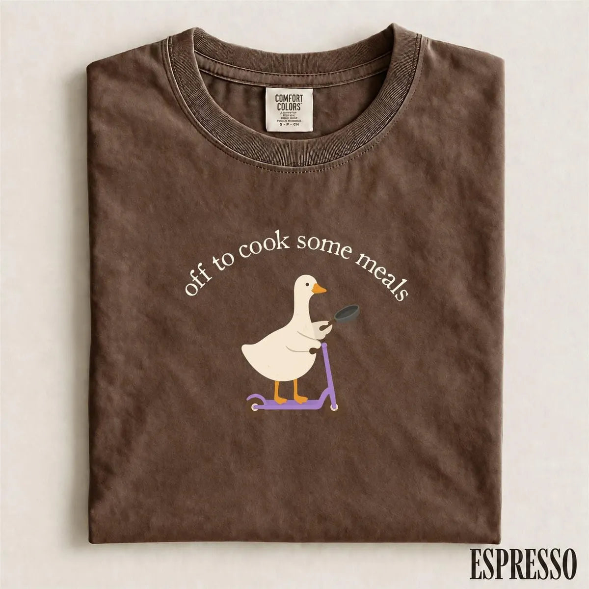 Folded Espresso brown Comfort Colors tee with a cream goose on a purple scooter holding a frying pan under arched text