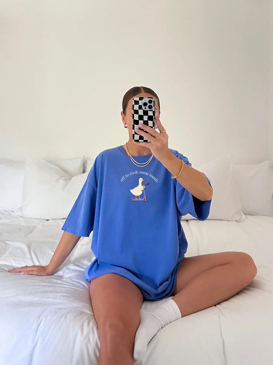 Woman in a Flo Blue Comfort Colors tee taking a mirror selfie on a bed, a small goose on a scooter under arched text