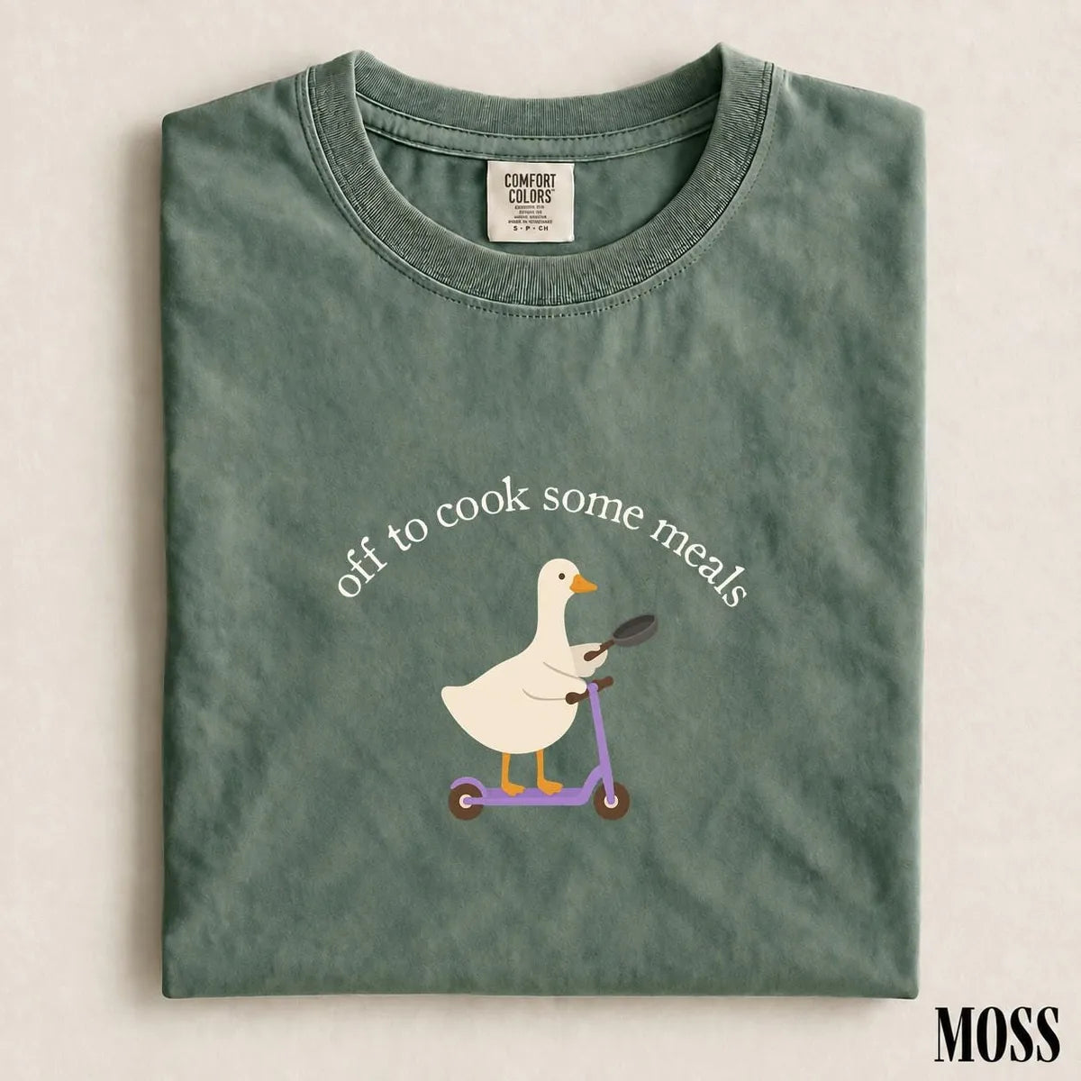 Folded Moss green Comfort Colors tee with a white goose holding a frying pan on a scooter under arched cook some meals