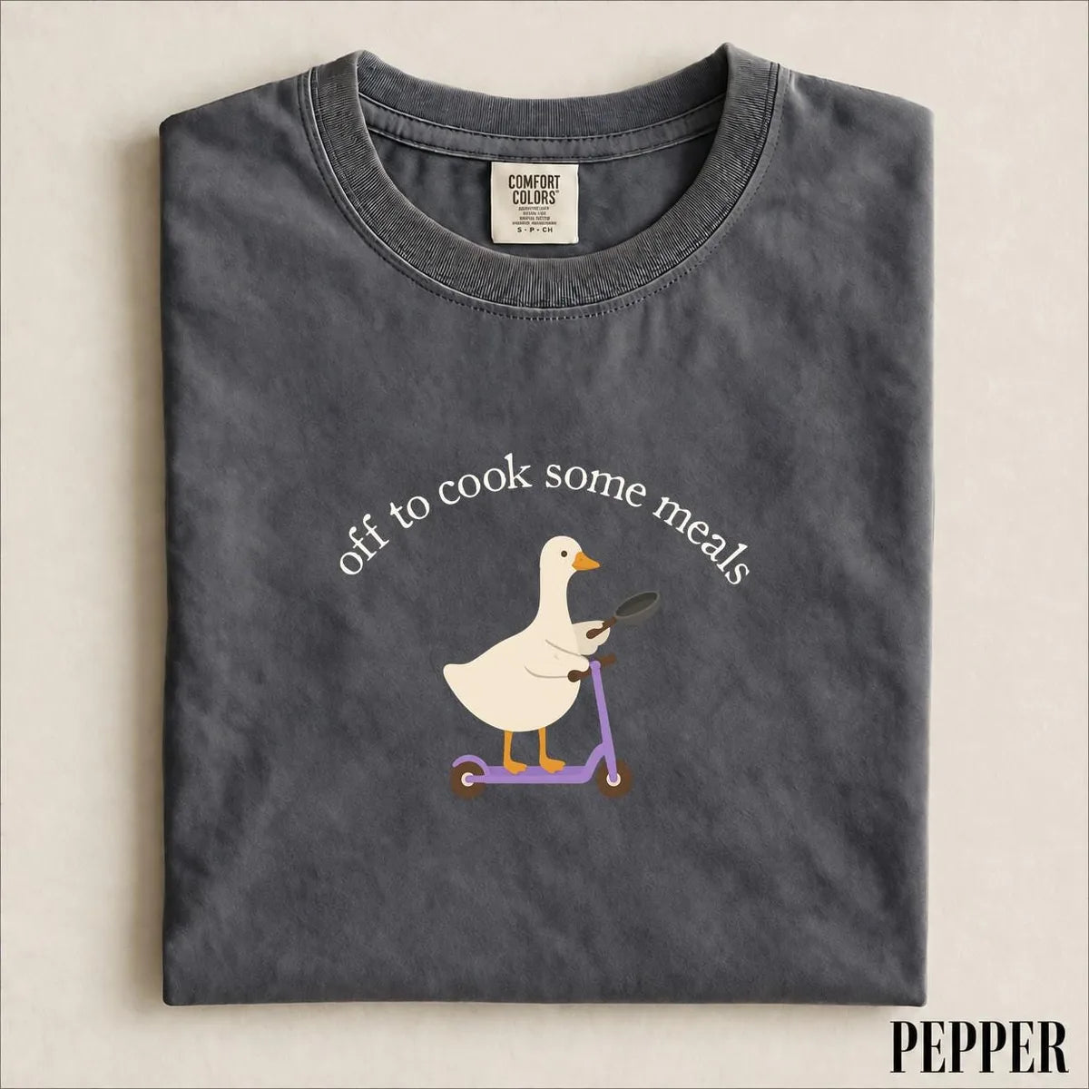Folded Pepper Comfort Colors tee with a goose holding a frying pan on a purple scooter under arched off to cook some meals