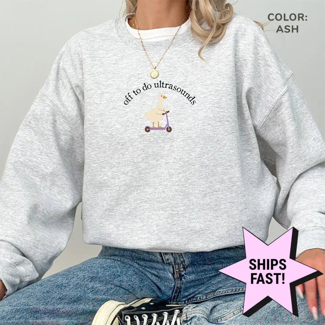 Woman in an Ash crewneck sweatshirt and jeans, a goose on a purple scooter under arched black text, off to do ultrasounds