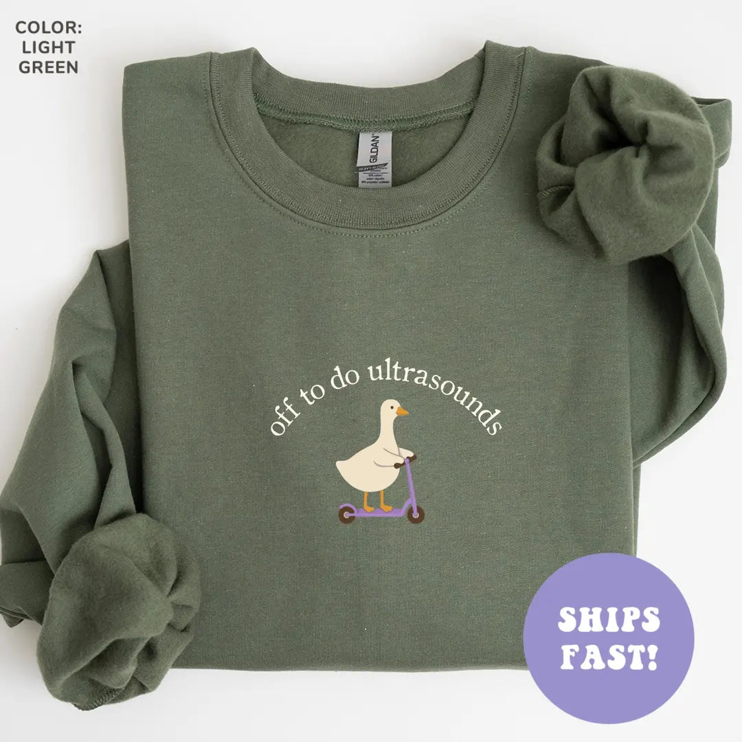 Flat lay Military Green sweatshirt with a goose on a purple scooter under arched cream text, off to do ultrasounds